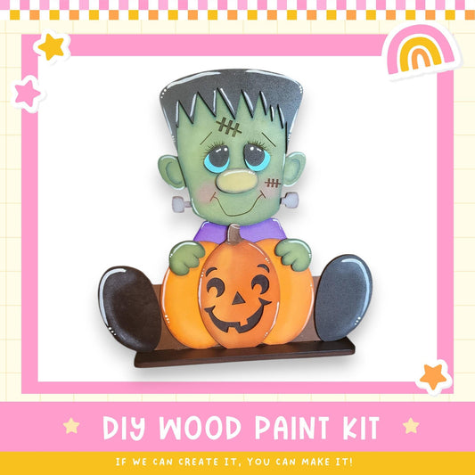 A DIY wood paint kit featuring a carved pumpkin and a carved green monster character, with a pink and yellow border and stars around the edges.