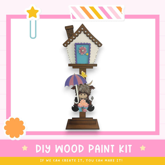 a DIY wood paint kit with a wooden house and a small cartoon character holding an umbrella. The kit appears to be made of wood and is designed for painting.