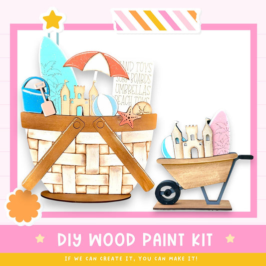 The image is a colorful and playful DIY wood paint kit, featuring a basket filled with various beach-themed items such as a surfboard, umbrella, sandcastle, and beach toys. The kit appears to be designed for children to create their own beach-themed artwork using the provided materials.