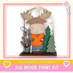 A wooden toy moose holding a fishing rod and surrounded by a Christmas tree and a snowman decoration, with a pink and yellow border and text promoting a DIY wood paint kit.