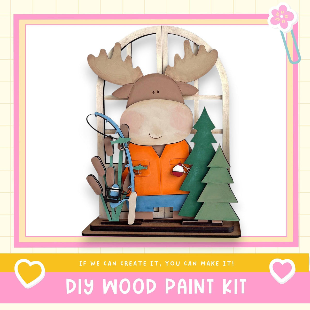 A wooden toy moose holding a fishing rod and surrounded by a Christmas tree and a snowman decoration, with a pink and yellow border and text promoting a DIY wood paint kit.