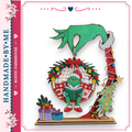 A hand-made ornament featuring a green Grinch-like character reading a book, surrounded by Christmas decorations such as a wreath, presents, and a Christmas tree.