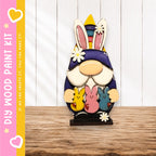 A wooden gnome figurine holding three colorful Easter bunny plush toys, set against a white wooden background.