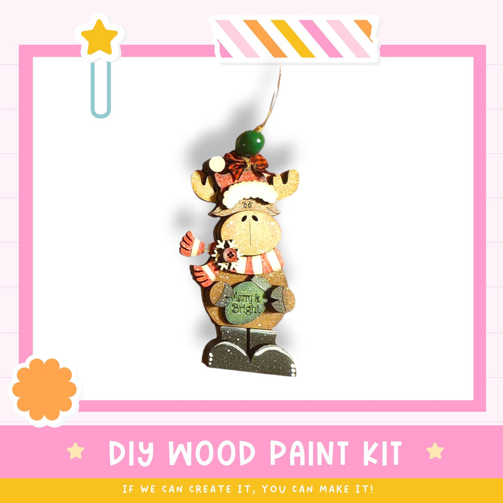 a wooden ornament in the shape of a moose, wearing a hat and scarf, with a green ball on its head. The image is part of a DIY wood paint kit, and the caption below the image encourages viewers to create their own wooden ornaments.