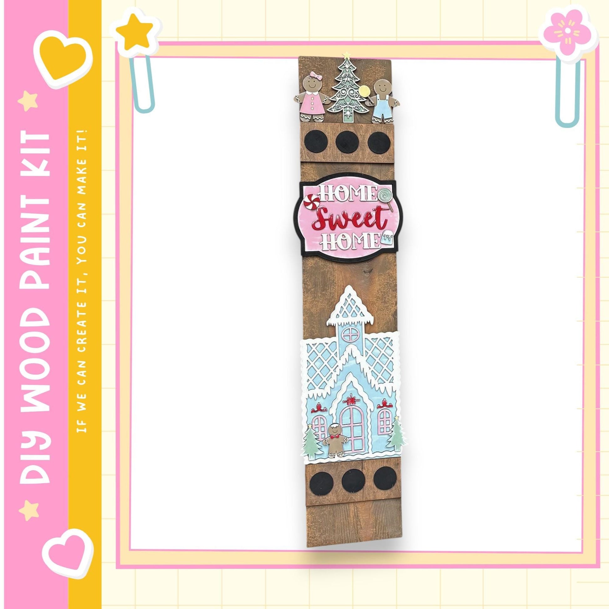 a wooden sign with a whimsical design, including a gingerbread house and other decorative elements. The sign is surrounded by a pink and yellow border, and there are small decorative elements such as hearts and stars on the border.