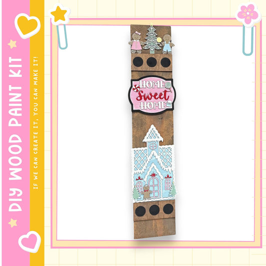 a wooden sign with a whimsical design, including a gingerbread house and other decorative elements. The sign is surrounded by a pink and yellow border, and there are small decorative elements such as hearts and stars on the border.