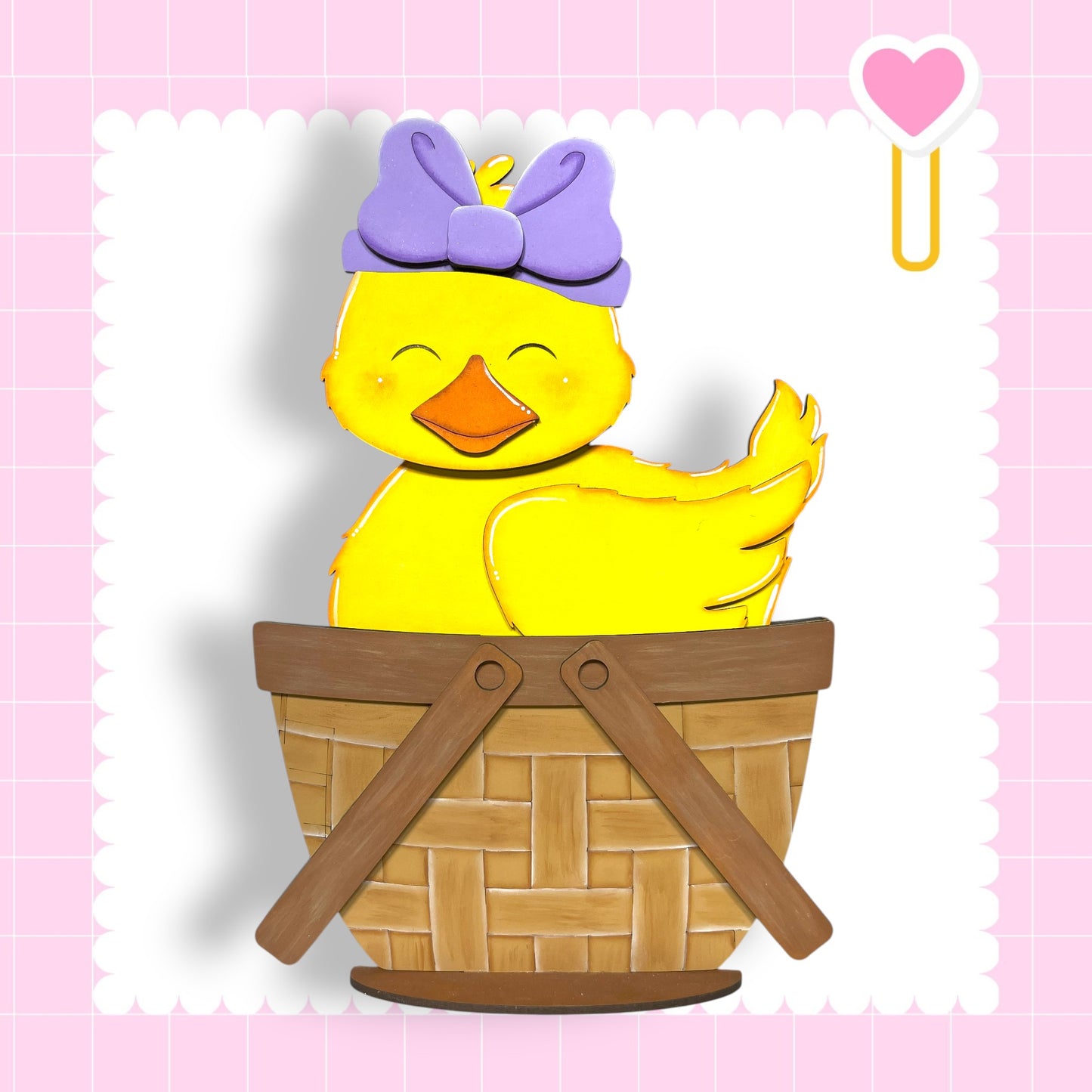 A yellow duck wearing a purple bow sits in a wooden basket against a pink background.