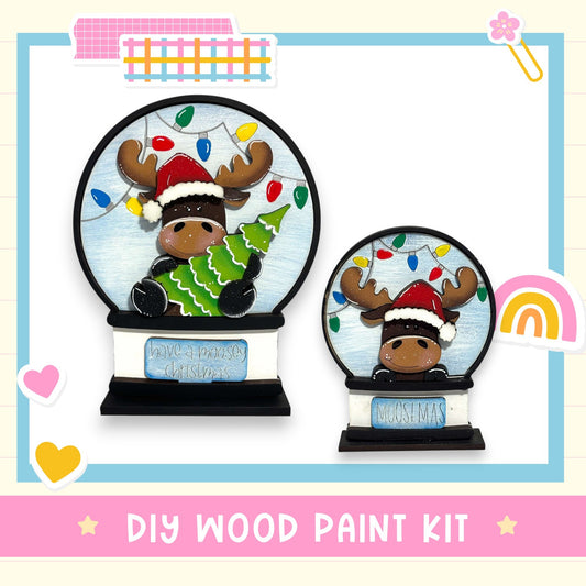 two snowglobe decorations, each with a moose wearing a Santa hat. The snowglobes are set against a background with Christmas lights and a rainbow. The image also includes text that reads "DIY WOOD PAINT KIT" and "HAVE A MOOSEY CHRISTMAS".