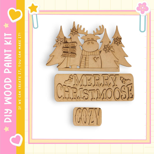 a wooden Christmas decoration kit with a moose and snowflakes. The kit includes a sign that says "MERRY CHRISTMOOSE" and a block that says "COZY". The kit is presented against a pink and yellow background with a heart and star design.