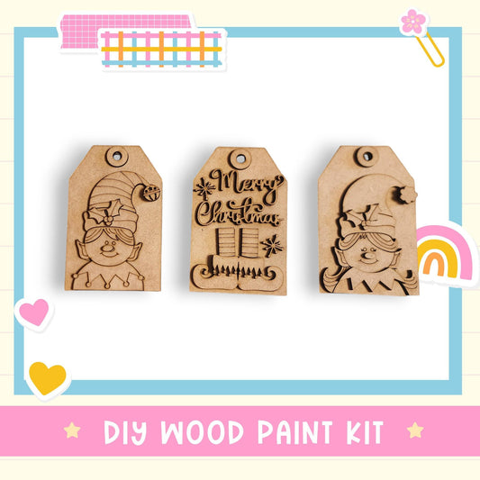 three wooden tags with festive designs, including a gnome and a Christmas tree, as well as a tag with the text "Merry Christmas". The tags are arranged in a row, and the background includes a pink and blue border with various colorful stickers. The image also includes the text "DIY WOOD PAINT KIT" at the bottom.