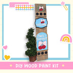The image is a colorful and whimsical DIY wood paint kit, featuring a wooden sign with a wreath, a heart, and a rainbow, along with a potted plant and other decorative elements.