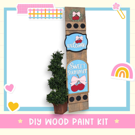 The image is a colorful and whimsical DIY wood paint kit, featuring a wooden sign with a wreath, a heart, and a rainbow, along with a potted plant and other decorative elements.