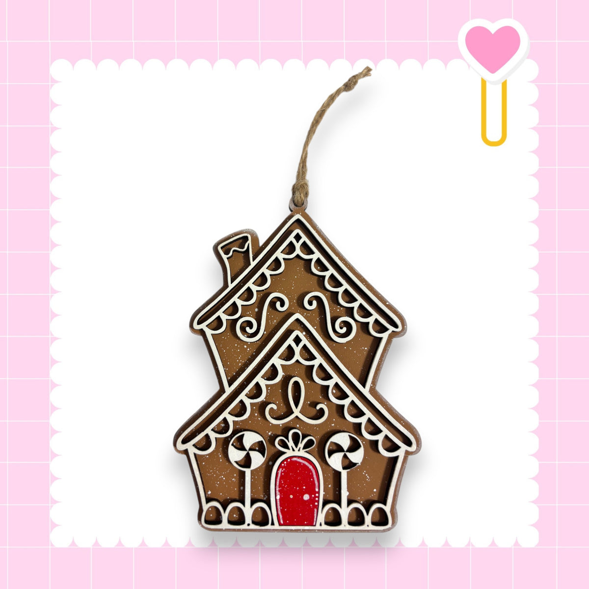 A decorative gingerbread house ornament with intricate white and brown designs, hanging from a string against a pink background with a scalloped edge.