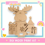 a DIY wood paint kit for creating a wooden Christmas tree decoration. The kit includes a wooden tree, a bow, and a star, all made of cardboard. The kit is presented in a pink and blue frame, with various decorative elements such as hearts and a rainbow.