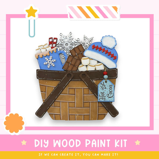 a DIY wood paint kit, which includes a wicker basket filled with various craft items such as a snowflake, marshmallows, and a blue hat. The kit appears to be made of wood and is designed for painting.