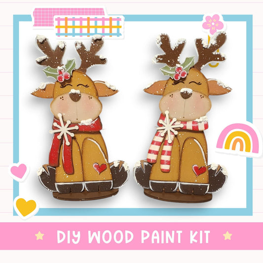 two wooden reindeer figures, each with antlers and a scarf, sitting on a white background. The reindeer figures are decorated with snow and flowers, and the image includes a DIY wood paint kit and a rainbow.