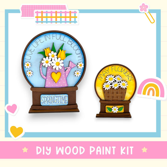 two wooden paint kits, one with a teapot and the other with a basket of flowers, both labeled "Springtime". The kits are presented against a white background with various decorative elements such as hearts and a rainbow. The text "DIY WOOD PAINT KIT" is displayed at the bottom of the image.