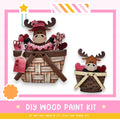two wooden moose figurines sitting in baskets, with hearts and other decorative elements. The image is a DIY wood paint kit, and it encourages viewers to create their own wooden moose figures using the provided kit.