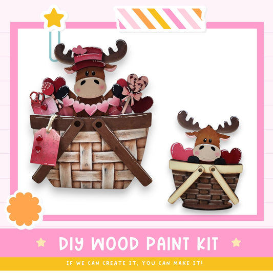 two wooden moose figurines sitting in baskets, with hearts and other decorative elements. The image is a DIY wood paint kit, and it encourages viewers to create their own wooden moose figures using the provided kit.