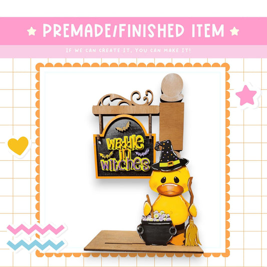 a yellow duck figurine dressed as a witch, holding a cauldron and standing next to a sign that says "Wackie in Witch's".