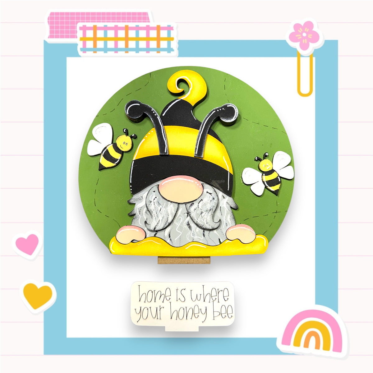 A gnome wearing a yellow hat and holding a honey bee is surrounded by a green circle, with two bees flying around it. The text "home is where your honey bee" is displayed below the gnome.
