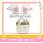 The image is a DIY wood paint kit featuring a basket filled with Easter eggs and a pair of bunny ears, set against a pink and white background.