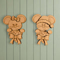 two wooden cutouts of cartoon mice, each wearing a bow and hat, hanging on a light green wooden wall.