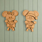two wooden cutouts of cartoon mice, each wearing a bow and hat, hanging on a light green wooden wall.