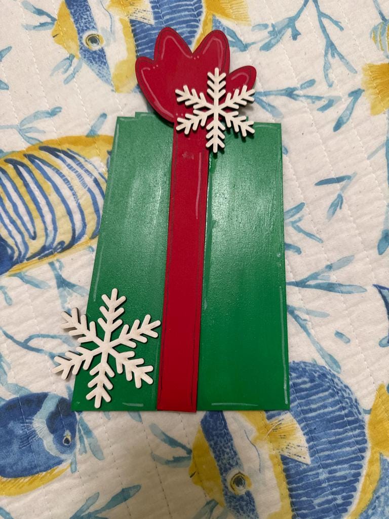 A green gift box with a red ribbon and two white snowflakes on top, placed on a patterned fabric background.