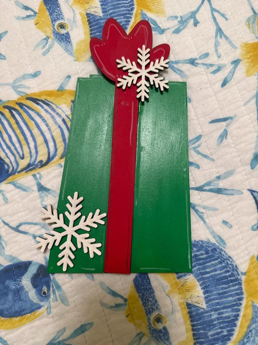 A green gift box with a red ribbon and two white snowflakes on top, placed on a patterned fabric background.