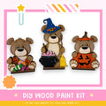 three wooden teddy bears, each dressed in a different costume, including a witch's hat, a pumpkin, and a pumpkin with a carved face. The bears are arranged in a row, with the witch's hat on the left, the pumpkin on the right, and the pumpkin with a carved face in the center. The image also includes text that reads "DIY WOOD PAINT KIT" and "IF WE CAN CREATE IT, YOU CAN MAKE IT!"