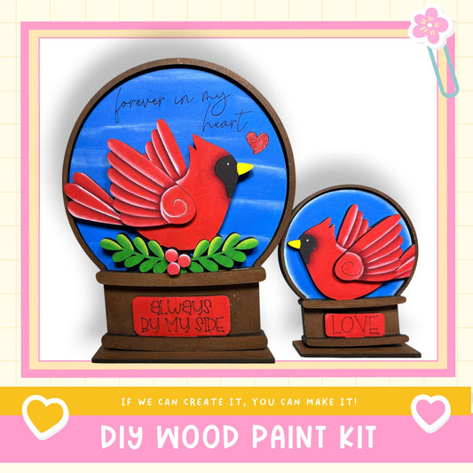 two wooden snow globes, each depicting a red cardinal bird. The snow globes are set against a blue background, and there is a message that reads "Forever in my heart" written on them. The image also includes a DIY wood paint kit, suggesting that the viewer can create their own snow globes.