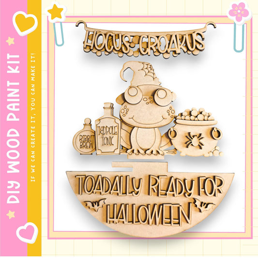 a Halloween-themed wooden craft kit, including a wooden frog, a wooden cauldron, and a wooden bottle labeled "Ribbit Brew". The kit is designed to be painted and used to create Halloween decorations.