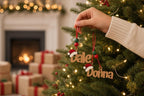 A hand is holding a pair of ornaments with the names "Dale" and "Donna" on them, hanging from a decorated Christmas tree.