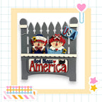 A decorative wooden sign with two cartoon-like figures, one wearing a blue shirt and the other a red shirt, sitting on a wooden bench. The sign has the text "God Bless America" written on it.