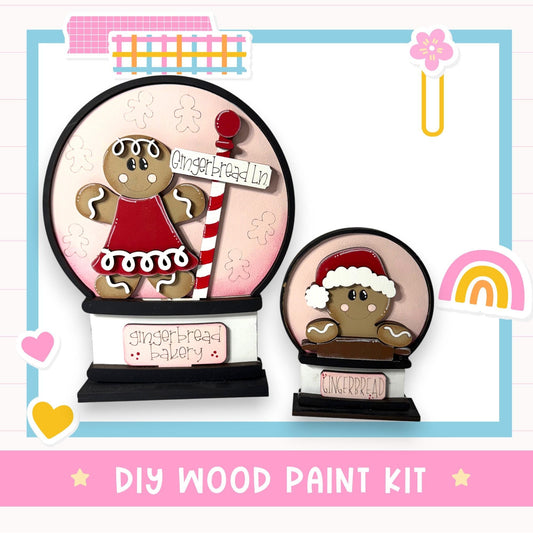 two wooden snow globes, one larger and one smaller, both decorated with gingerbread men and labeled "Gingerbread Bakery". The larger snow globe is placed on a black base, while the smaller one is on a pink base. The image also includes various decorative elements such as a heart, a rainbow, and a flower. The text "DIY WOOD PAINT KIT" is displayed at the bottom of the image.