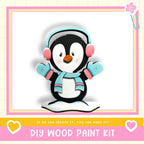 A wooden penguin figurine wearing headphones and a scarf, with the text "DIY WOOD PAINT KIT" and "If we can create it, you can make it!"
