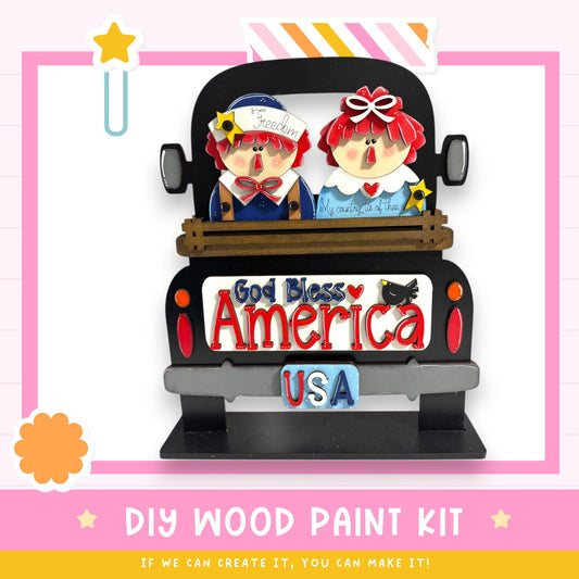 A DIY wood paint kit featuring a wooden truck with two cartoon characters, one wearing a blue hat and the other a red hat, sitting in the back. The truck has a "America" license plate and the text "God Bless America USA" is displayed on the back.