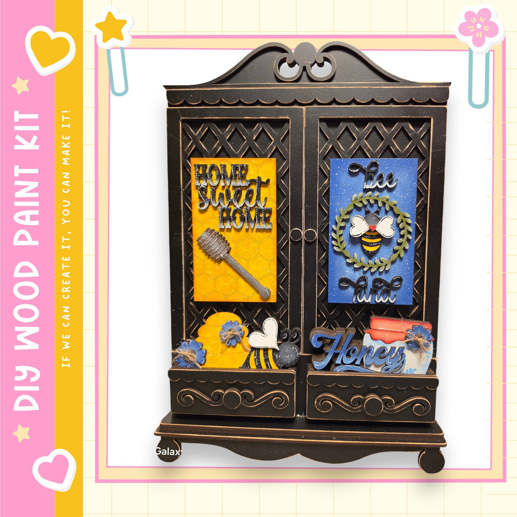 The image is a wooden paint kit featuring a decorative cabinet with various items, including a bee, a honey jar, and a wreath.