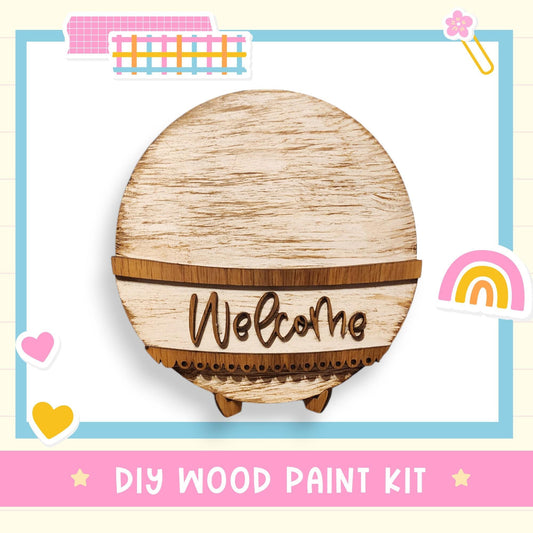 a DIY wood paint kit with a wooden sign that says "Welcome" and various colorful stickers surrounding it.