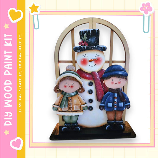 A wooden snowman with a carrot nose and coal eyes stands on a wooden base, accompanied by two children dressed in winter clothing.