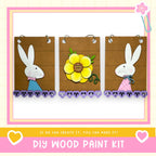 The image is a DIY wood paint kit featuring three wooden panels with illustrations of a white rabbit and a yellow flower. The panels are hanging on a wall, and the image includes text that encourages creating art with the kit.