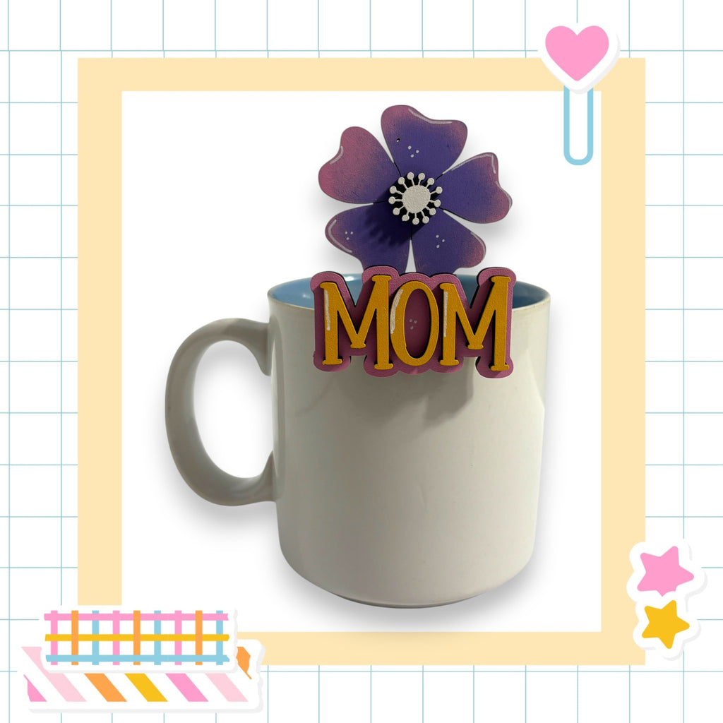 A white mug with the word "MOM" written on it, placed on a table surrounded by various colorful and decorative elements.