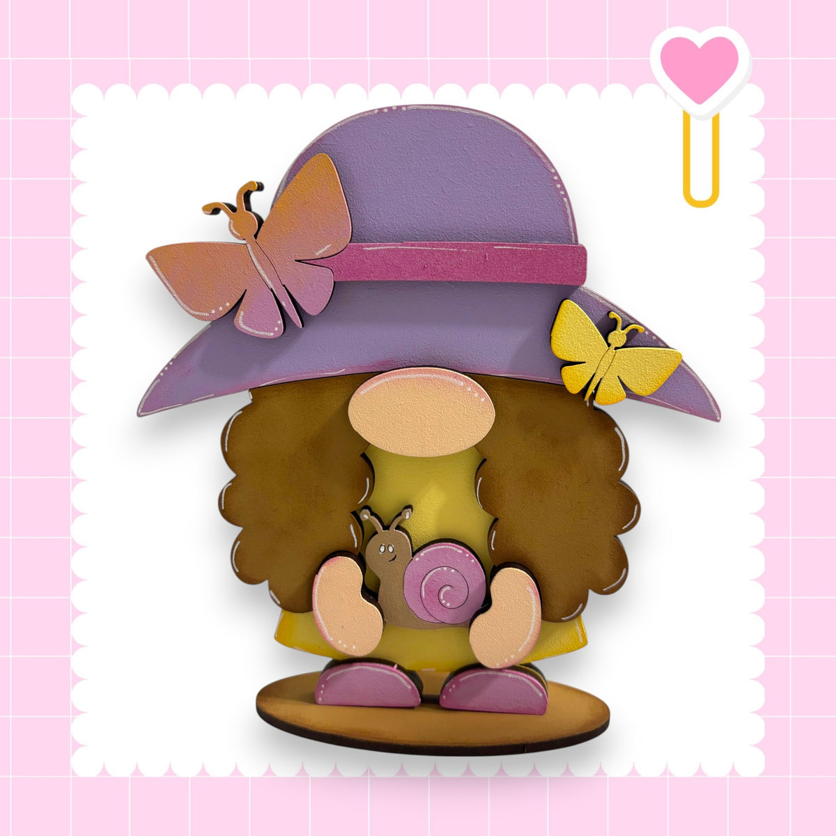 A whimsical wooden gnome figure wearing a purple hat with a pink ribbon and holding a snail, set against a pink background with a heart shape.