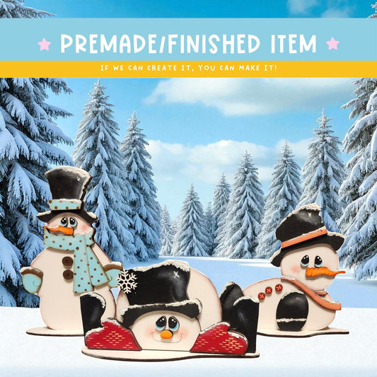 three snowman decorations against a snowy winter landscape with pine trees in the background.