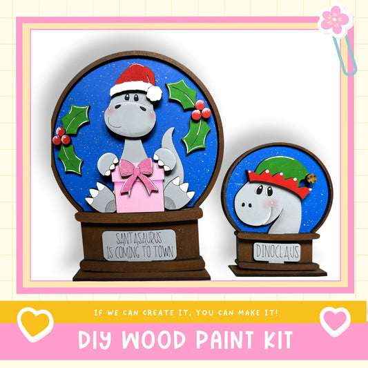 two wooden snow globes, one larger and one smaller, decorated with festive elements such as holly leaves and a Santa Claus hat. The larger snow globe is labeled "Santasaurus is coming to town" and the smaller one is labeled "Dinoc Claus". The image also includes text that says "If we can create it, you can make it!" and "DIY WOOD PAINT KIT".