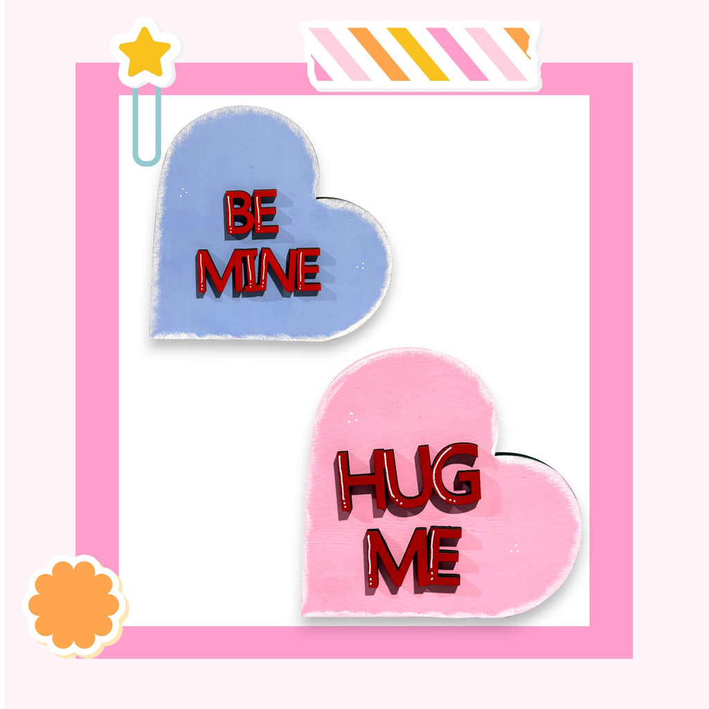 two heart-shaped objects with the words "Be Mine" and "HUG ME" written on them, set against a pink background.