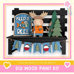 A DIY wood paint kit featuring a moose figure holding a fishing rod, surrounded by a "Keep it reel" sign, a bench, and a garland with Christmas trees.