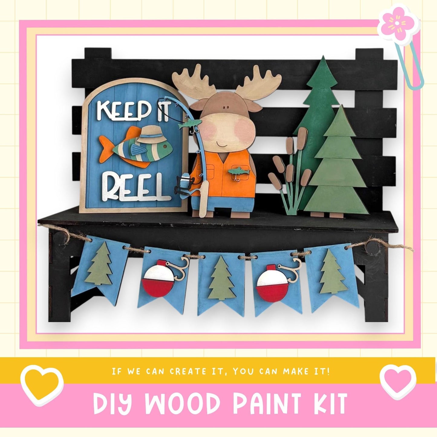 A DIY wood paint kit featuring a moose figure holding a fishing rod, surrounded by a "Keep it reel" sign, a bench, and a garland with Christmas trees.
