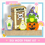 The image is a DIY wood paint kit featuring a frog, a pumpkin, and various Halloween-themed items such as a spell book and a poison bottle.
